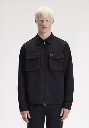 Jeune personne à la peau claire et aux cheveux clairs portant une veste noire zippée avec deux poches à rabat à l'avant, debout devant un fond uni.