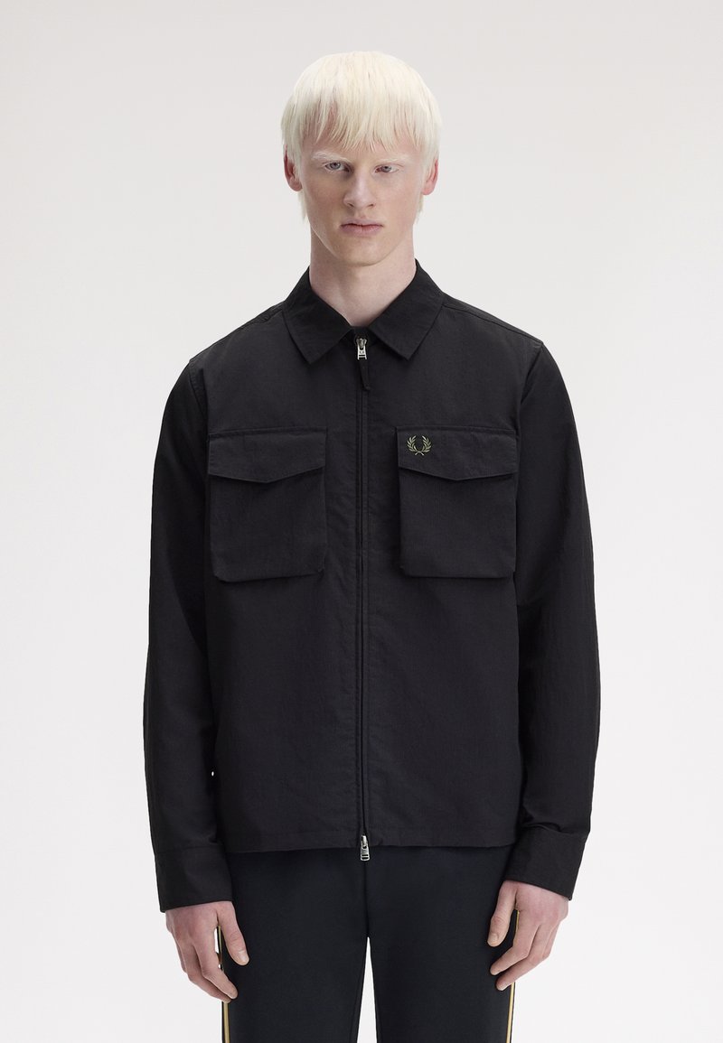 Persona joven con piel clara y cabello claro que lleva una chaqueta negra con cremallera y dos bolsillos delanteros con solapa, de pie frente a un fondo liso.