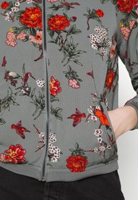 Kwiatowy rozpinany hoodie z teksturowanej szarej tkaniny, z wyrazistymi czerwonymi i różowymi kwiatami, zielonymi liśćmi oraz małymi ptakami. Zawiera boczne kieszenie.