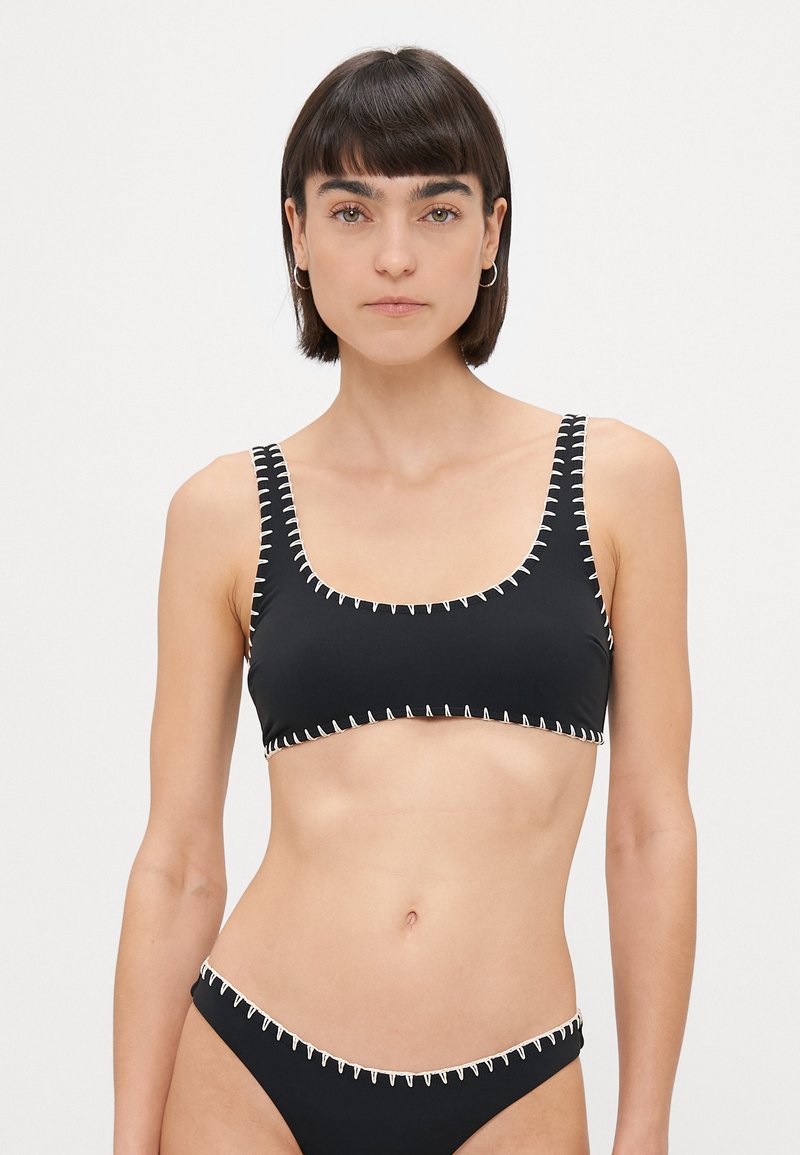 Svart bikini med firkantet utringning og brede stropper, med kontrasterende hvite sømmer langs kantene. Jevn tekstur, minimalistisk design.