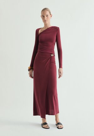 Mujer con vestido burdeos de manga larga y un solo hombro, con detalle fruncido en un lateral y sandalias planas negras, de pie frente a un fondo uni.
