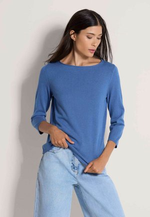 Vrouw met lang donker haar die een blauw shirt met driekwart mouwen en lichtblauwe jeans draagt, poseert met haar handen dichtbij haar zakken tegen een effen achtergrond.
