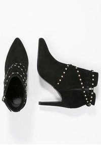 Botines de ante negro hasta el tobillo con punta afilada, tacones de aguja y tachuelas doradas a lo largo de las correas. Presentan una textura suave y un diseño entrelazado.