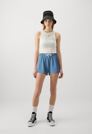 Shorts en coton bleu clair avec une taille élastique et un cordon blanc, associés à un débardeur crème et un chapeau seau noir. Baskets montantes noires.