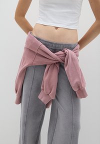 Helles rosa Sweatshirt, das um die Taille gebunden ist, über grauen, weiten Sweatpants. Weicher Stoff mit gerippten Bündchen und lässiger Passform.
