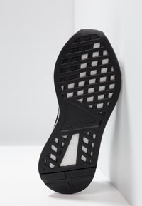 Svart gummisula till sneakers med ett texturerat mönster, som har geometriska utskärningar och Adidas-logotypen. Står upprätt mot en vit bakgrund.