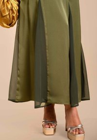 Gonna verde oliva con pannelli trasparenti, abbinata a sandali platform argentati. Il tessuto ha una consistenza liscia e una leggera lucentezza, creando profondità.