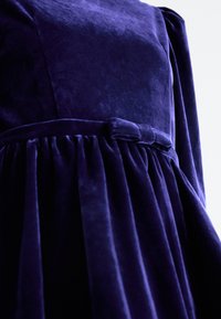 Violet samettikjole med en tætsiddende overdel, indsamlet skirt og en bælteliv med en sløjfe detaljer. Glat tekstur og rig farve.