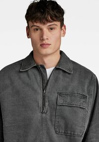 Siva denim jakna z zadrgo spredaj, ovratnikom in žepom na prsih; strukturirana zasnova z gladko teksturo.