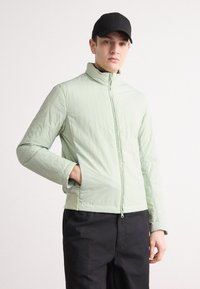 Jachetă verde deschis, cu fermoar, guler înalt și buzunare laterale, cu o suprafață texturată și un design ajustat. Asortată cu pantaloni negri.