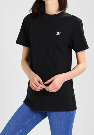 Person iført en sort Adidas t-shirt med logo på brystet, kombineret med blå jeans, hænder løst foldet foran.
