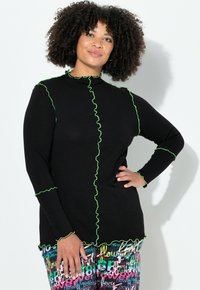 Zwarte top met lange mouwen en groene geplooide stiksels langs de hals, naden en zoom. Gecombineerd met kleurrijke broek met patroon en tekstontwerpen.