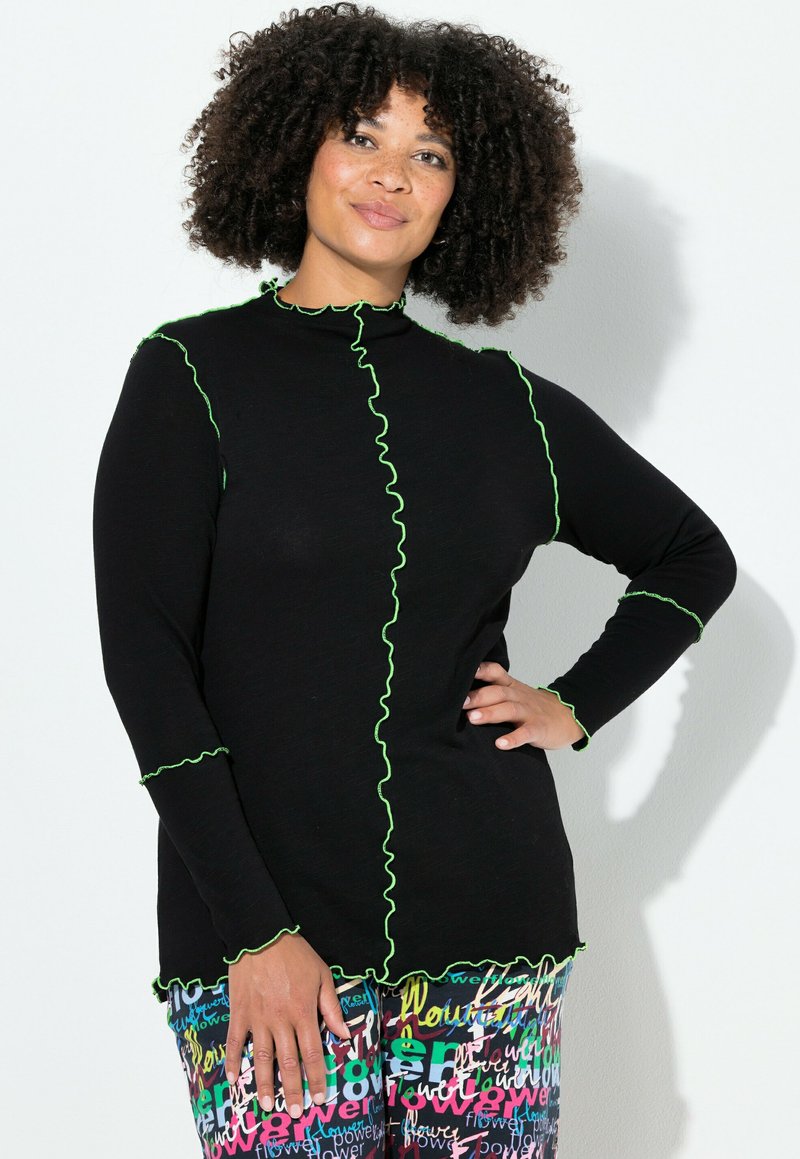 Zwarte top met lange mouwen en groene geplooide stiksels langs de hals, naden en zoom. Gecombineerd met kleurrijke broek met patroon en tekstontwerpen.