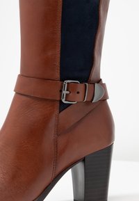 Bota de tacón de cuero marrón con panel de ante azul marino, detalle de hebilla en el tobillo, textura suave y diseño de tacón cuadrado.