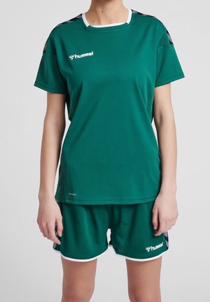 Person iført grønt hummel sportstøjssæt med kortærmet skjorte og shorts, med sorte chevron skulderaccents og hvid kant.