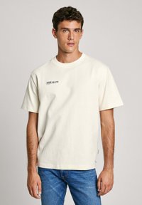 Camiseta de algodón color crema con mangas cortas y escote redondeado, que presenta un pequeño logo en navy en el pecho y un corte relajado.
