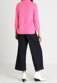 Měkký růžový svetr s tříčtvrtečními rukávy a bočními rozparky, v kombinaci s tmavými širokými džínami a bílými kotníkovými botami.