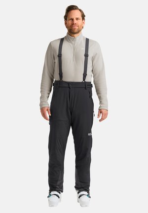 Pantalons de neige noirs avec bretelles réglables, poches zippées et un logo. Portés par-dessus un polaire gris clair avec un col à demi-zip.