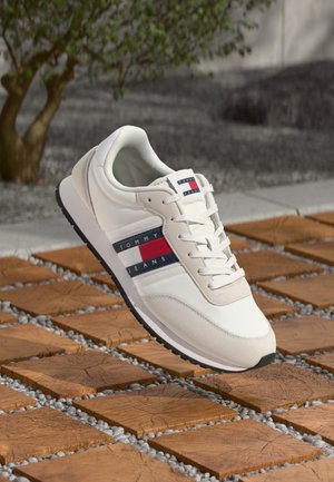 Baskets blanches avec des matériaux en daim et en cuir, affichant un logo Tommy Jeans rouge et bleu audacieux, un design à lacets et une semelle en caoutchouc noire.