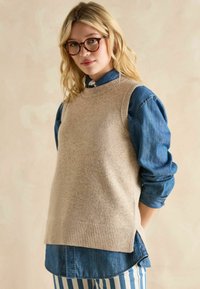 Beige gestrickte Weste mit geripptem Saum, getragen über einem dunkelblauen Denimhemd. Der Hintergrund hat eine sanfte, hell-neutrale Farbe.