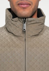 Geacă puf maro deschis cu model texturat în formă de diamant, guler înalt și închidere cu fermoar prevăzută cu accesorii argintii.