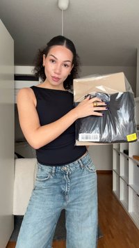 Czarny element odzieży zapakowany w plastik, trzymany przed osobą ubraną w czarny bezrękawnik i jasnoniebieskie dżinsy, w pomieszczeniu.