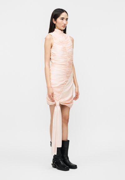 Νέα γυναίκα με ροζ αμάνικο φόρεμα με ρυτίδες και μακρύ ζωνάρι δεμένο στο τελείωμα, φορώντας μαύρες μπότες μέχρι τη μέση της γάμπας, στέκεται μπροστά σε απλό λευκό φόντο.