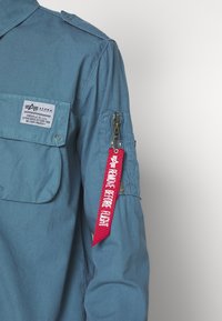 Manche de veste bleu de style militaire avec une poche zippée dotée d'une étiquette en tissu rouge "Remove Before Flight" et d'une étiquette Alpha Industries.