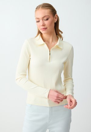 Kobieta o jasnych włosach, ubrana w kremowy, prążkowany, długi rękaw polo z półzamkiem, poprawiająca mankiet, na jednolitym białym tle.