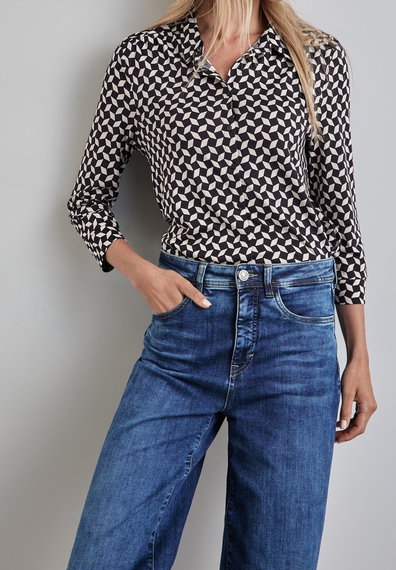 Femme portant une chemise boutonnée à motif géométrique noir et blanc avec des manches trois-quarts et un jean bleu en denim taille haute, main dans la poche.