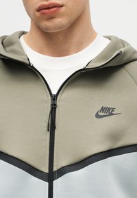 Vyras dėvi spalvų blokų Nike megztinį su priekiu užsegamu užtrauktuku, alyvuogių žalios viršutinę dalį, šviesiai mėlyną apatinę dalį ir juodą logotipą ant krūtinės