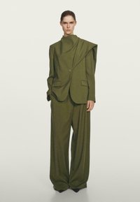 Costume taillé vert olive avec un blazer structuré à large col et fermeture à un bouton, associé à un pantalon ample.