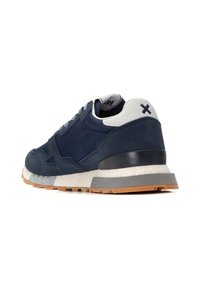 Zapatilla azul marino hecha de materiales lisos y de ante, con una pestaña en el talón blanca, suela gris y suela de goma texturizada.