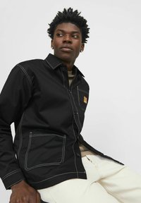 Black button-up jacket with white stitching and a chest pocket; features a collar and a small logo patch. Worn over a beige shirt.