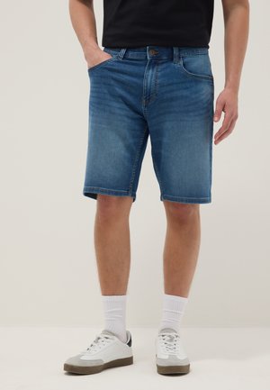 Homme portant un short en jean bleu moyen, des chaussettes blanches, des baskets blanches et une chemise noire, debout avec une main dans la poche.