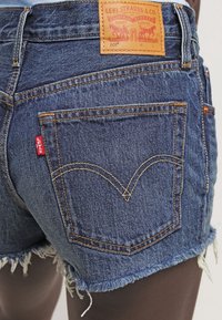 Jeansshorts med fransad nederkant, mörkblå färg och synlig sömnad. Har en lädermärke tagg och en röd logotyp på bakfickan.