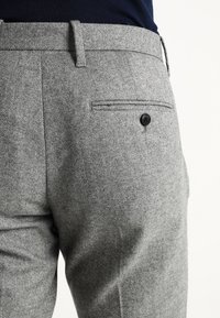 Grijze visgraatbroek met een gladde textuur, voorzien van een achterzak met een zwarte knoop en een op maat gemaakte tailleband.