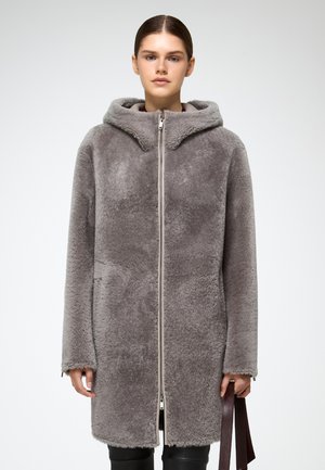 Kvinde iført en lang grå faux fur frakke med hætte og lynlås fortil, som holder mørkebrune stropper i højre hånd.