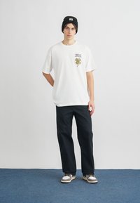 Biała bawełniana koszulka z haftowanym wzorem koniczyny w kolorze zielonym i brązowym. Luźne czarne spodnie i czarna czapka. Szaro-białe sneakersy.