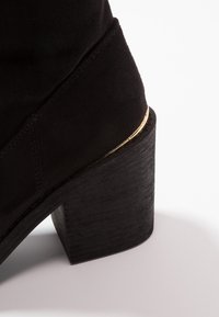 Bottine en daim noir avec talon bloc et détail de bordure dorée entre le talon et la tige, posée sur une surface blanche.