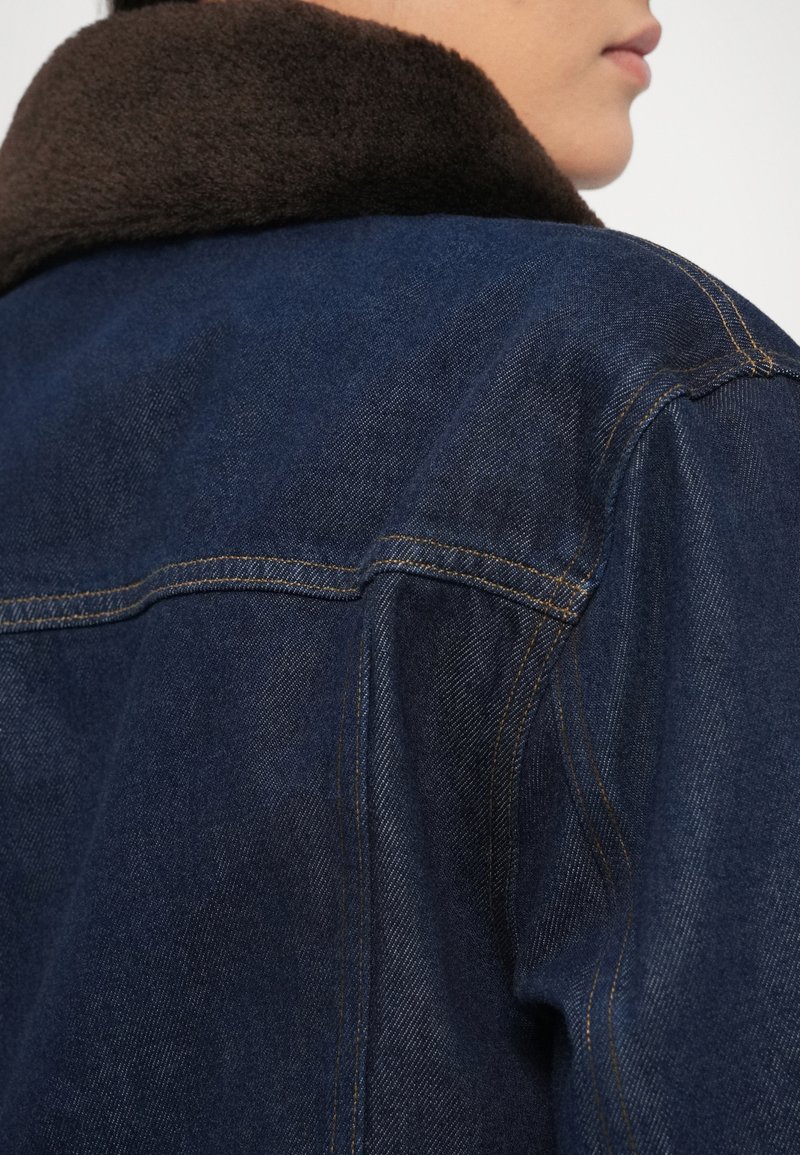 Nahaufnahme einer Person, die eine dunkelblaue Jeansjacke mit braunem Fleece-Kragen trägt, Rückansicht der Schulter und teilweise Profil sichtbar.