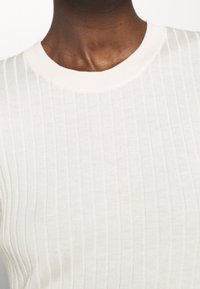 Bílý žebrovaný pletený top s kulatým výstřihem a mírně přiléhavým střihem, zdůrazňující hebkou texturu a jemné horizontální pruhy.