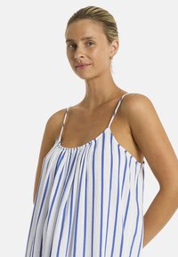 Una donna con un'espressione serena indossa un vestito senza maniche a righe con linee verticali blu e bianche, girata leggermente di lato.
