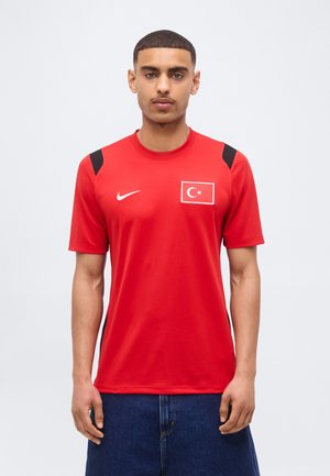 Mężczyzna w czerwonej sportowej koszulce z krótkim rękawem, z emblematem tureckiej flagi i logo Nike, w połączeniu z ciemnoniebieskimi dżinsami.