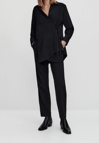 Mustapaitainen pitkähihaisella kääntöpaidalla ja sidotulla vyöllä, yhdistetty suoralahkeisiin mustiin housuihin. Sileä kankaan rakenne, nilkkurit täydentävät asukokonaisuuden.