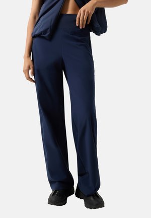 Person trägt dunkelblaue weite Hose und schwarze Schuhe, hebt dabei leicht das Oberteil an, um die Taille zu zeigen.