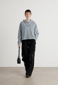 Lys grå cropped hettegenser med brodert logo, kombinert med svarte cargo bukser med flere lommer. Modellen bærer en svart håndveske.