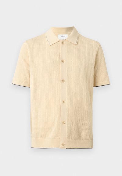 Μπεζ πλεκτό πουκάμισο με κοντά μανίκια, κουμπιά μπροστά, μυτερό γιακά, υφή υφάσματος και διακριτική σκούρα διακόσμηση στις μανσέτες των μανικιών και το τελείωμα.