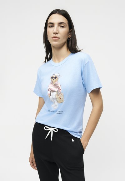 Γυναίκα με μακριά σκούρα μαλλιά φοράει ανοιχτό μπλε μπλουζάκι με μια αρκούδα με γυαλιά ηλίου και ριγέ πουκάμισο, συνδυασμένο με μαύρο παντελόνι με κορδόνι.