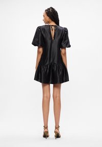 Μαύρο φόρεμα φτιαγμένο από λεία ύλη, με φουσκωτά μανίκια, ρυθμισμένο στρίφωμα και πίσω άνοιγμα σε σχήμα κλειδαρότρυπας με δέσιμο. Φορεμένο με ψηλές γόβες.